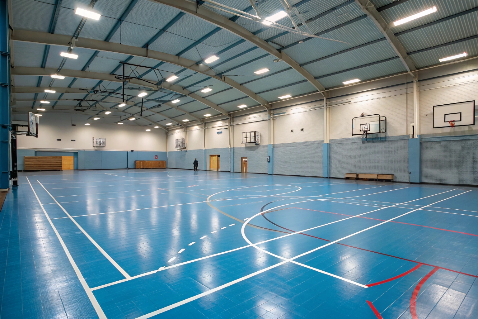 indoor sports hall with blue epoxy floor coating, white court line markings visible, basketball hoops on walls, bright overhead lighting illuminating the space